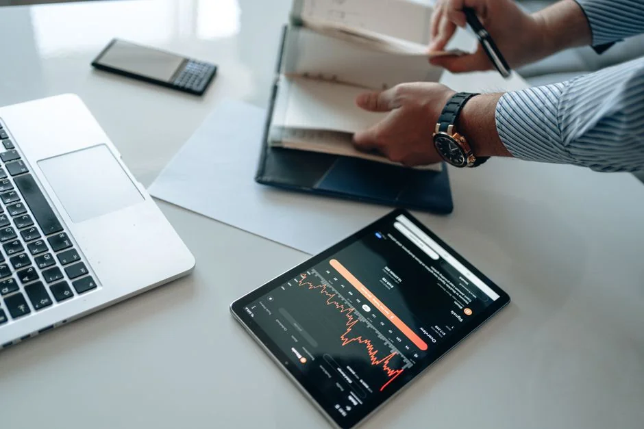 Hands using smartphone beside laptop with stock charts, showcasing digital trading.